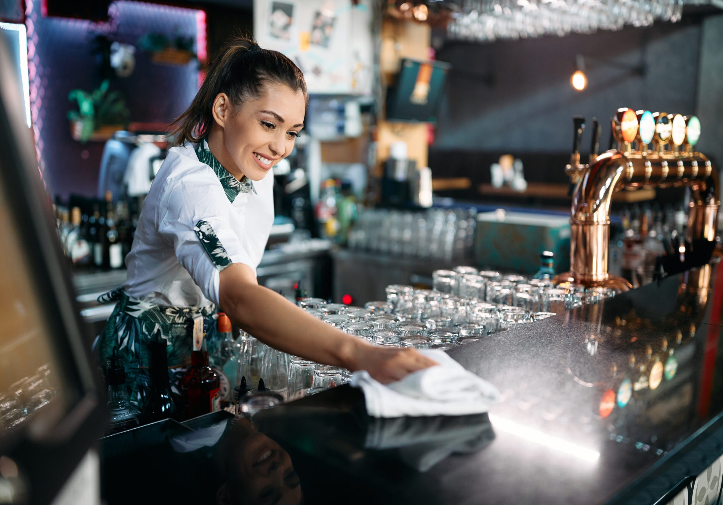 Happy female bartender wipes bar counter after working hours.